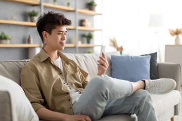 Smiling asian man using cellphone at home sitting on couch