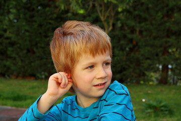 The blond boy looks into the distance with interest, pinching his ear. A five-year-old boy looks away with a smile.