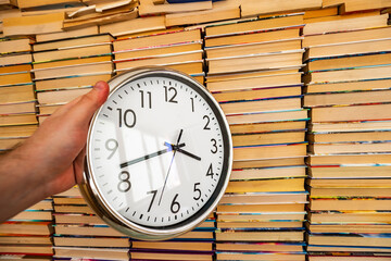 Many books in stacks and white big round clock keep in hand on the books background. Time and books that can be read for life. Many multicolored books in columns next to the clock in hand