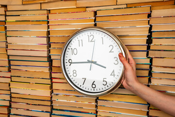 Many books in stacks and white big round clock keep in hand on the books background. Time and books that can be read for life. Many multicolored books in columns next to the clock in hand