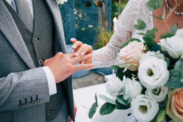 Wedding ceremony of dressing gold rings with a bouquet of white and pink flowers in the hands of the bride in a white dress and the groom in a gray suit on the festive site