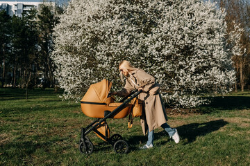 Blonde woman walking with a stroller in a park, talking with her baby.