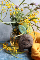 Cute crochet toy, acacia flowers in a glass jar close up photo. Grey textured background with copy space. Handmade gift ideas.