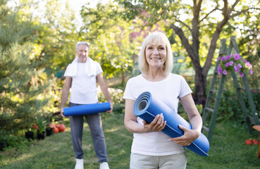 Obraz premium Portrait of active senior woman posing with husband in garden, holding yoga mats, exercising together outdoors