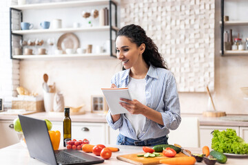 Positive woman writing new recipe while watching video on laptop, cooking in modern kitchen interior