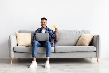Smiling man using pc and credit card sitting on couch