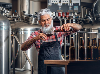 Shot of bearded senior brewer pouring his product to glass around table with beer and barrels.