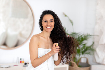 Smiling young caucasian brunette lady in towel applies spray oil on long curly hair for treatment in bedroom interior - obrazy, fototapety, plakaty