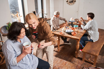 Women drinking tea from the ceramic cups and smiling while looking at the smartphone