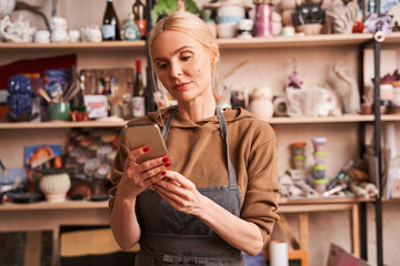 Woman wearing apron relaxing at her art studio and reading messages at the smartphone
