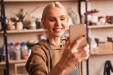 Cheerful female artist holding mobile phone and making self portrait on it at her art studio