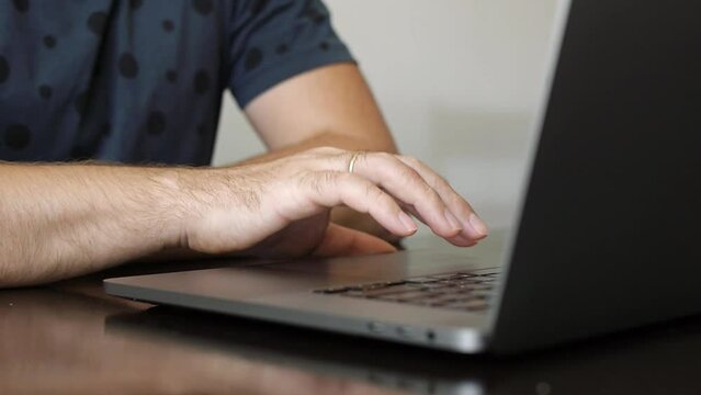 Close-up of male hands scrolling touchpad of silver laptop. A man wearing casual t-shirt works on a laptop in a cafe or coworking space. Slow motion