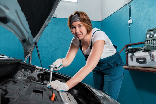 Smiling mechanic woman in blue coveralls repair a engine with ratchet wrench. The concept of woman's equality and works at auto repair shop