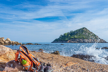 Backpack and hiking shoes on empty beach in Argentario region, Tuscany, Italy. Sand bay in natural park, island in mediterranean sea blue waving water - obrazy, fototapety, plakaty