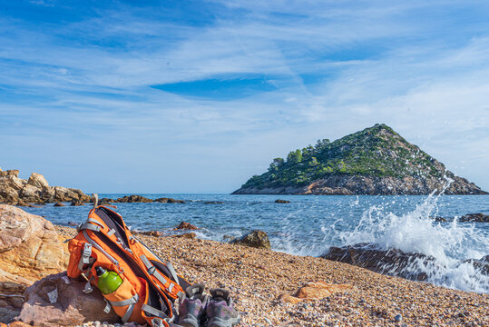 Backpack and hiking shoes on empty beach in Argentario region, Tuscany, Italy. Sand bay in natural park, island in mediterranean sea blue waving water