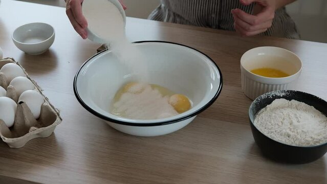 Woman adding sugar in a bowl with eggs to beat the dough. The process of making dough for buns with ingredients on a table.