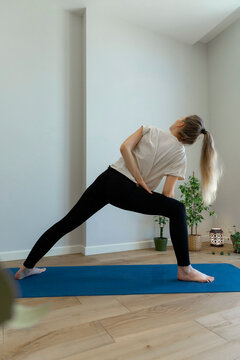 flexible young woman is doing standing twisting pose, stretching her shoulder and back, wearing beige t-shirt, black leggings and ponytail on blue aport matt at her home 