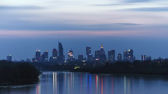 Time-lapse evening to night transition in big city of Warsaw skyline, high skyscrapers illumination reflected on Wisla river surface. Downtown beautiful cityscape panorama lit with warm sunset light