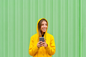 Panoramic front view of cheerful woman holding a phone on green background. Horizontal view of woman with yellow raincoat using technology isolated on green wall. People and technology concept.