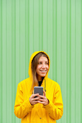 Close-up side portrait of blonde woman wearing a raincoat. Horizontal view of caucasian woman in the street with yellow raincoat isolated on green wall. People isolated in background with copy space.