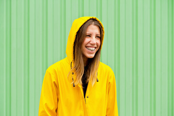 Middle angle portrait of happy woman wearing a raincoat. Horizontal view of caucasian woman in the street with yellow raincoat isolated on green wall. People isolated in background with copy space.