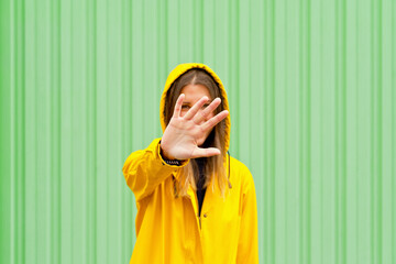 Mid waist view of woman covering her face in a yellow raincoat. Horizontal view of caucasian woman outdoors with yellow raincoat isolated on green wall. People isolated in background with copy space.