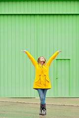Vertical front view of woman raising arms in a yellow raincoat standing on the street. Full length body of woman under the rain isolated on green wall. People isolated in background concept.