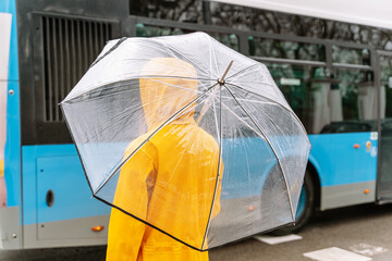 Close-up of unrecognizable woman with umbrella waiting for the bus. Horizontal rear view of woman at bus stop with transparent umbrella outdoors. Public transport and weather concept