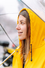 Vertical side view of woman at traffic light with transparent umbrella outdoors. Close-up view of woman walking in yellow raincoat under rain with umbrella. Weather concept