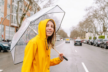 Horizontal front view of woman crossing the street with transparent umbrella outdoors. Mid waist view of woman walking in yellow raincoat under rain with umbrella. Weather concept