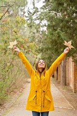 Front view of happy woman with raincoat raising arms with maple tree leaves. Vertical mid waist view of woman looking up at fallen leaves in yellow hoodie outdoors. People and nature backgrounds.