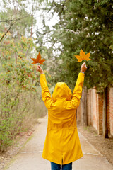 Rear view of unrecognizable woman raising arms with maple tree leaves. Vertical mid waist view of woman looking at fallen leaves in yellow raincoat outdoors. People and nature backgrounds.