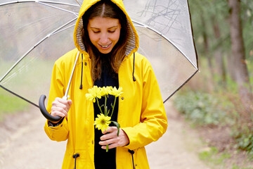 Front view of happy woman walking with umbrella and flowers in park. Horizontal mid waist view of woman walking in park with yellow raincoat and daisies bouquet. People and nature
