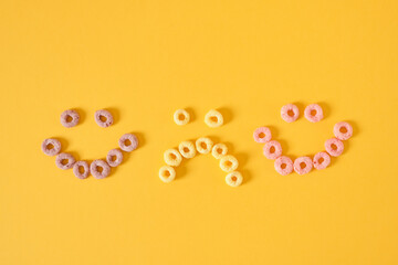 colored breakfast cereals laid out in the shape of a smiley face on a yellow background top view