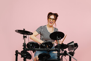 Energetic eccentric lady playing on electric drums. Her hair in buns. Over pink background. Grimacing, looking in the camera.