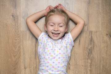 Fototapeta premium Smiling child girl with closed eyes lying on a wooden floor. Top view, flat lay.