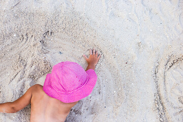 Child girl in pink panama hat is played with sand on the beach. Top view, flat lay.
