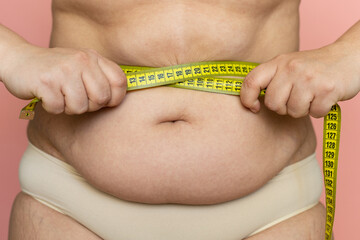 Cropped photo of naked fat woman tummy, trying to lose weight. Holding and measuring by roulette tape belly, abdomen. Large waist, big size xl. Health care, obesity and overweight problem. Close-up. 