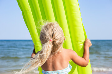 Blond child girl in a swimsuit holding an inflatable mattress on the beach.
