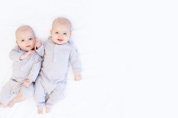 two newborn baby twin girls in a cotton suit on a white bed at home lie and smile, a place for text