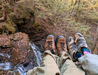 Man and woman hiking boots feet setting on overlook bluff looking at waterfalls in late summer in the forest and wearing brown hiking pants.