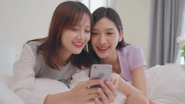 Young beauty Asian women LGBT lesbian happy couple laying on the bed using smartphone together in bedroom. LGBT lesbian couple together indoors concept. Spending nice time at home