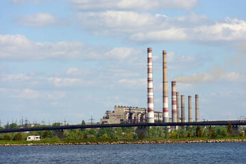 Many smokestack are behind river on sky with clouds background.