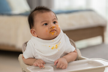 Baby face dirty after feeding pumpkin mashed enjoy and amazed.Adorable smudgy asian baby learning to eating and feeding vegetable mashed happiness at home.