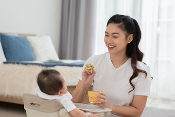 Obraz na płótnie Canvas Mother feeding her asian baby daughter with pumpkin mashed or vegetable mash for vitamin on rubber spoon.Mom trying feed little baby boy at home enjoy and spending time together.Baby feeding Concept