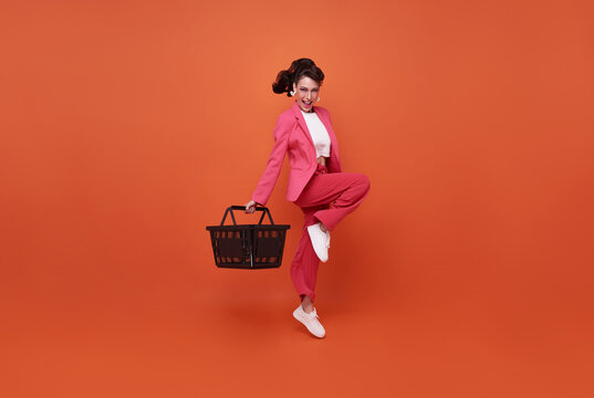 Smiling happy woman holding shopping basket and jumping isolated on orange background.