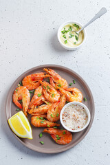 Shrimps cooked with spices lie on a plate with lemon, salt, herbs and sauce on a gray background.