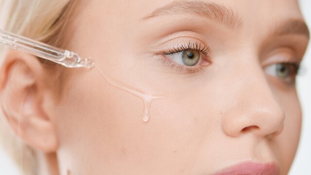 Extreme close-up shot of good-looking Caucasian young blond woman puts skin care serum on her cheek from dropper on white background | Skin care serum advertising