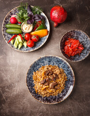 Pilaf with lamb meat in large dish, plate of tomato salad and dish with vegetables and greens