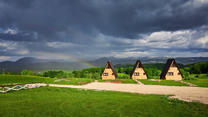 Landscape with rainbow over wooden cabins. A frame wooden houses in countryside with dramatic sky....